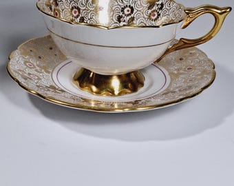 May include: A white teacup and saucer with a gold and pink floral pattern. The cup has a scalloped rim and a gold handle. The saucer has a gold rim and a pink floral pattern.