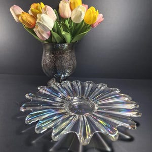 May include: A clear, iridescent glass serving dish with a scalloped edge, resembling a flower, sits in front of a decorative glass vase filled with colorful tulips. The tulips are a mix of yellow, white, pink, and orange.