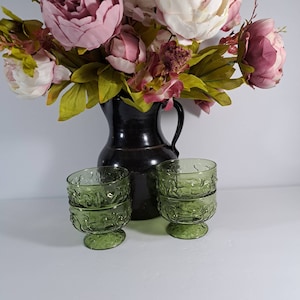 May include: Four green glass dessert dishes with a textured design. The dishes have a footed base and are arranged in front of a black ceramic jug.