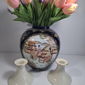 May include: Two white ceramic candle holders with gold trim and a detailed leaf design. The candle holders are in front of a blue and white porcelain vase with a Japanese scene painted on it. The vase is filled with pink, white, and purple tulips.