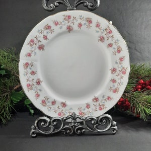 Peut inclure: Une assiette à dîner en porcelaine blanche avec un motif floral délicat de fleurs roses et bleues sur une vigne. L'assiette a un bord doré et est exposée sur un support en métal noir.