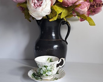 May include: A white teacup and saucer with a floral design featuring white flowers and green leaves. The teacup and saucer are sitting in front of a black pitcher with a bouquet of pink and white flowers.