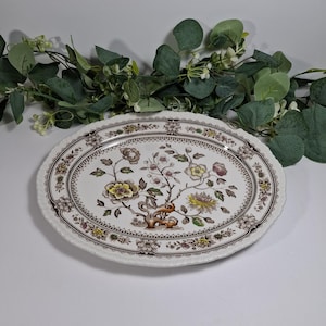 May include: An oval serving platter with a floral design. The white platter features a brown and yellow floral pattern with a scalloped edge. Greenery is in the background.