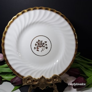 May include: White ceramic plate with a gold rim and a gold floral design in the centre. The plate is on a gold metal stand.