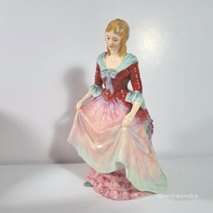 May include: A porcelain figurine of a woman in a long, flowing gown. The dress is primarily red, with a pink and light blue skirt. The figurine has blonde hair and is holding up the skirt of her dress.