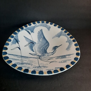 May include: A blue and white ceramic plate featuring a hand-painted design of a bird in flight. The bird is blue and white with yellow accents. The plate has a yellow and blue border.