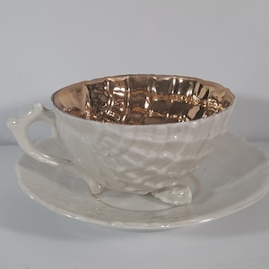 May include: A white teacup and saucer set with a gold interior. The cup has a textured exterior and a unique, sculpted handle. The saucer has a simple, elegant design.