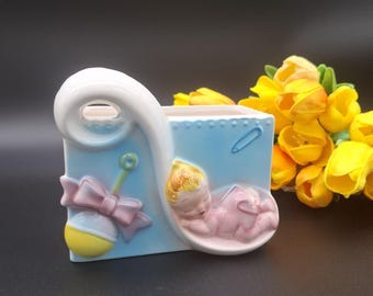 May include: A light blue ceramic planter featuring a sculpted sleeping baby in a pink cradle, a yellow and white rattle with a pink bow, and a blue safety pin. The planter has a white curved handle and a smooth, glossy finish.