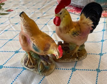 May include: Two ceramic rooster figurines. One is a hen, the other a rooster. The rooster has a red comb and wattle, with yellow, brown, and black feathers. The hen is brown and orange. Both are on a base with green and brown accents.