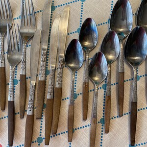 May include: A collection of vintage silverware, including forks, knives, and spoons. The silverware features a decorative floral design on the handles and a two-tone color scheme of silver and brown. The utensils are arranged on a patterned tablecloth.