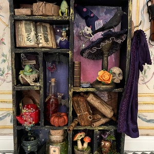 Pode incluir: Uma caixa de sombra de madeira preta cheia de itens temáticos de Halloween, incluindo um chapéu de bruxa, um caldeirão, uma garrafa de poção, uma abóbora e um corvo. A caixa é dividida em compartimentos e tem um acabamento desgastado.
