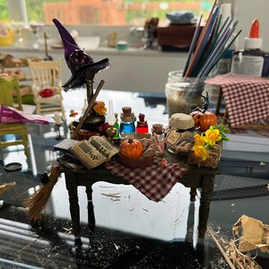 Pode incluir: Uma mesa de bruxa em miniatura com um chapéu roxo, um livro, uma vassoura e várias garrafas de poções. A mesa é decorada com uma toalha de mesa xadrez vermelha e branca e uma pequena abóbora.
