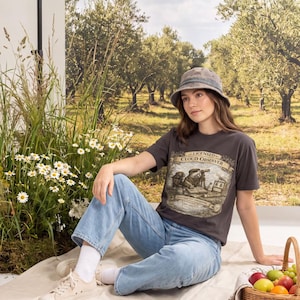 Könnte beinhalten: Ein anthrazitfarbenes Kurzarm-T-Shirt mit einem Grafikdruck einer Kreatur, die ein Teleskop benutzt, und dem Text "LICENSED CLOUD OBSERVER," kombiniert mit einer hellblauen Jeans, einem grauen Fischerhut, weißen Socken und beigen Stoffschuhen.