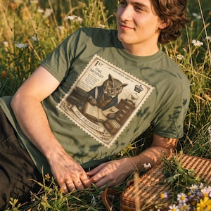 May include: An olive green t-shirt featuring a graphic print of an owl dressed in a waistcoat and bowtie, sitting at a desk with books and a typewriter. The design is framed like a postage stamp with the text 1ST OWL ARCHIVIST and VICTORIAN JOOS.