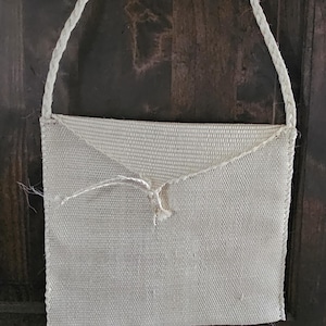 May include: A handmade, off-white woven bag with a braided rope strap. The bag has a flap closure secured with a knotted string. The texture of the bag is visible, suggesting a natural fiber material.