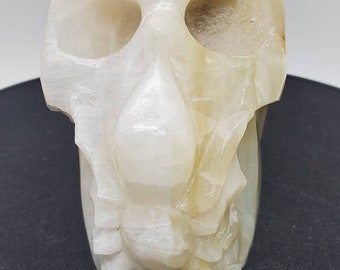 May include: A white alabaster skull sculpture with intricate details. The skull is carved with a realistic appearance, including eye sockets, a nose, and a jawline.