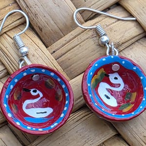 May include: A pair of drop earrings featuring small, bowl-shaped charms painted in a vibrant red with a white swan illustration in the center. The rims are accented with a bright blue border and white dots, suspended from silver-toned metal hooks.