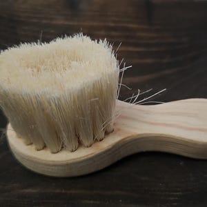 Puede incluir: Un pequeño cepillo de madera con cerdas naturales. El cepillo tiene una forma redondeada y un mango de madera.