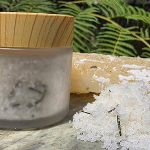 Op de afbeelding: Een matglazen pot met een houten deksel, een stuk zeep en een hoop witte badzouten met groene accenten. De pot en zeep zijn licht van kleur en de achtergrond heeft groen gebladerte.