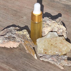 May include: A small glass bottle with a gold cap and dropper filled with a clear yellow liquid. The bottle is surrounded by several pieces of light brown, yellow, and white resin.