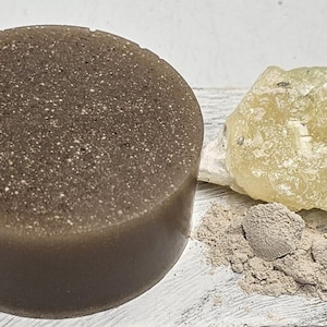 May include: A round, brown soap bar with a speckled texture, a piece of yellow-white resin, and a pile of light brown powder on a white surface.