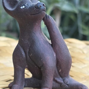 Könnte beinhalten: Eine dunkelbraune Keramikfigur mit matter Oberfläche, die eine Katze in aufrechter Sitzposition darstellt. Die Katze ist mit einem zum Kopf erhobenen Hinterbein dargestellt, als würde sie sich am Ohr kratzen, mit spitzen Ohren und kleinen Augen.