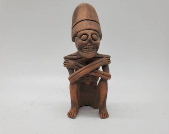 May include: A terracotta sculpture of a seated figure with a skull-like face and crossed arms. The figure is wearing a hat and has a detailed, carved design.
