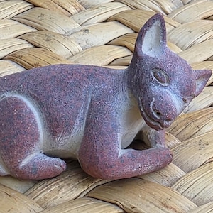 May include: A small, hand-carved stone figurine of a feline creature in a resting position. The sculpture features a mottled reddish-brown exterior with a smooth, matte texture, pointed ears, and carved facial details including eyes and a mouth.