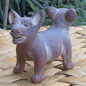 May include: A small, reddish-brown clay figurine of a dog with pointed ears, a wide grin showing teeth, and a short, upright tail. The surface of the sculpture has a textured, speckled appearance with lighter beige accents on the ears and mouth.