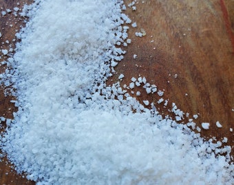 May include: Close-up of three piles of white salt crystals on a wooden surface. The salt varies in texture, with some appearing coarse and others finer. The wooden surface has a warm, brown tone, providing a contrast to the bright white salt.