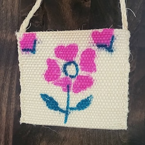 May include: A small, white, woven bag with a pink flower design. The flower has a blue center and green leaves. The bag has a rope handle.