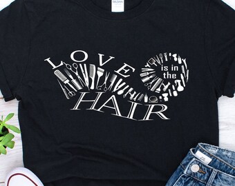May include: Black t-shirt with the text "LOVE is in the HAIR" in white, featuring a design of hair styling tools. The shirt is paired with blue jeans and white sneakers, suggesting a casual, everyday style.
