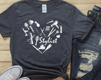 May include: A dark gray t-shirt with a white heart design featuring various hair styling tools and the word "Stylist". The shirt is paired with blue jeans and white sneakers.