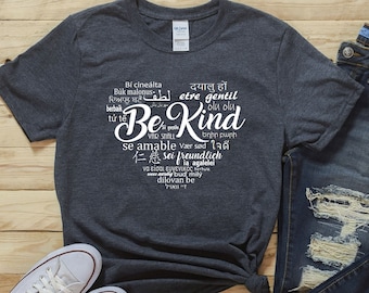 May include: A dark gray t-shirt with a white graphic design that says "Be Kind" in multiple languages. The shirt is laid flat on a wooden surface with a pair of white sneakers and a pair of blue jeans with distressed details.