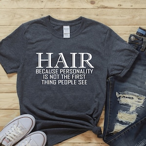 May include: A dark gray t-shirt with white text that reads "HAIR because personality is not the first thing people see". The shirt is paired with blue jeans and white sneakers.