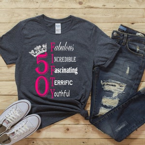 May include: A dark grey t-shirt with a pink number 50 and a silver crown. The text "Fabulous, Incredible, Fascinating, Terrific, Youthful" is written vertically next to the number.