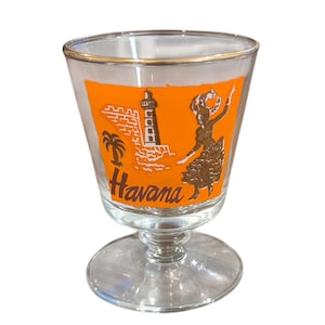 May include: Clear glass shot glass with a gold rim and a stem. The glass features an orange background with a white illustration of a lighthouse, palm tree, and a dancing figure. The word "Havana" is printed in white.