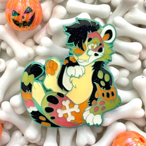 May include: A colorful enamel pin featuring a cartoon feline character with a green, black, and orange design, surrounded by orange pumpkin beads and white bone-shaped beads. The pin has a light blue border and a cross design.