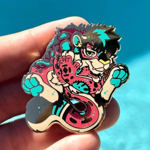May include: A colorful enamel pin featuring a stylized feline character with a snake-like tail. The pin has a black, teal, pink, and white color scheme. The character is wearing glasses and has the word "EAT" on its chest.