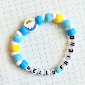 May include: A beaded bracelet with a mix of blue, white, and yellow beads. The bracelet has the words "HELLO FRIEND" spelled out in white letter beads. A round charm with a cartoon design is also included.
