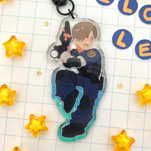 May include: A clear acrylic keychain featuring a cartoon illustration of a character with light brown hair, wearing a dark blue tactical uniform with a badge, holding a black handgun, attached to a black metal lobster clasp and ring.