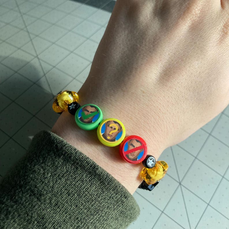 May include: A beaded bracelet featuring round beads with images of a man's face and other symbols. The bracelet includes yellow and black beads and is worn on the wrist.