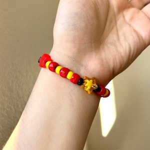 May include: A beaded bracelet with red, yellow, and black beads. A translucent yellow star charm is attached. The bracelet is worn on a wrist.