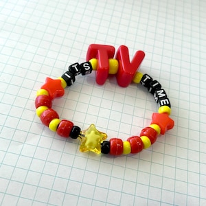May include: A vibrant beaded bracelet spelling "TV TIME". The bracelet is made of red, yellow, black, and clear beads. It includes star-shaped and round beads. The letters "TV" are red, and the text "ITS TIME" is spelled out.