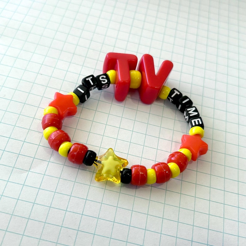 May include: A vibrant beaded bracelet spelling "TV TIME". The bracelet is made of red, yellow, black, and clear beads. It includes star-shaped and round beads. The letters "TV" are red, and the text "ITS TIME" is spelled out.