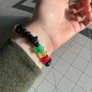 May include: A beaded bracelet featuring black star-shaped beads, interspersed with green, yellow, and red star beads. The bracelet is worn on a wrist, showcasing the colorful design and playful aesthetic. The bracelet is a fun accessory.