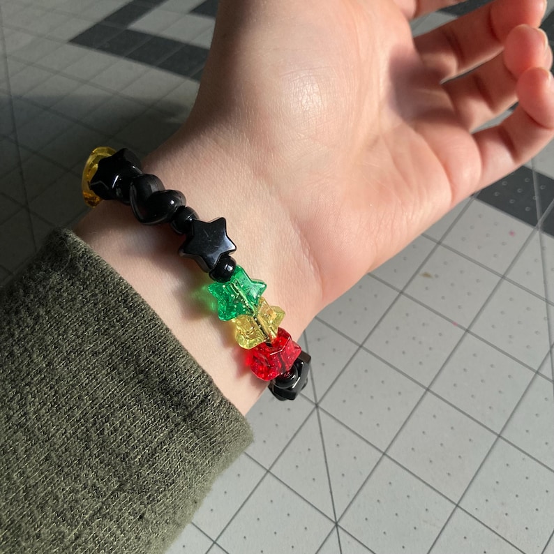 May include: A beaded bracelet featuring black star-shaped beads, interspersed with green, yellow, and red star beads. The bracelet is worn on a wrist, showcasing the colorful design and playful aesthetic. The bracelet is a fun accessory.