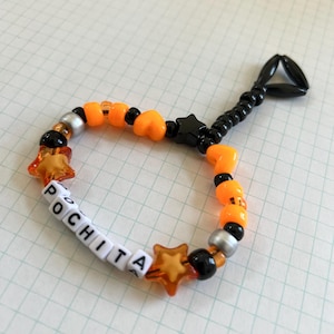 May include: A beaded bracelet with the name "POCHITA" spelled out in white letter beads. The bracelet features orange, black, and silver beads, along with star and heart-shaped accents. A black beaded loop is attached.