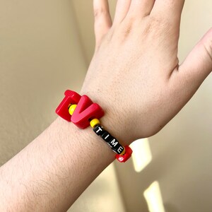 May include: A red bracelet featuring the word "TIME" in black and white beads. The letters "TV" are in red with yellow detailing. A vibrant and playful wrist accessory.