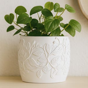 Puede incluir: Una maceta de cerámica blanca con un diseño de hojas y vides en relieve, llena de una planta verde frondosa. La maceta tiene forma cilíndrica y se asienta sobre una superficie de color claro. Las hojas de la planta tienen forma de corazón.